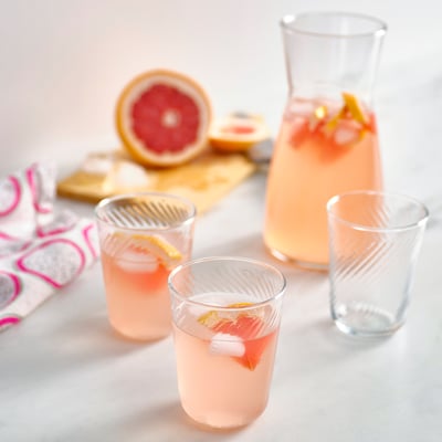 KLIPPLAX glazen set: drie roze drankjes in dunne, geribbelde glazen naast een grotere kan, gehalveerde grapefruit en servetten met patronen op een houten plank.