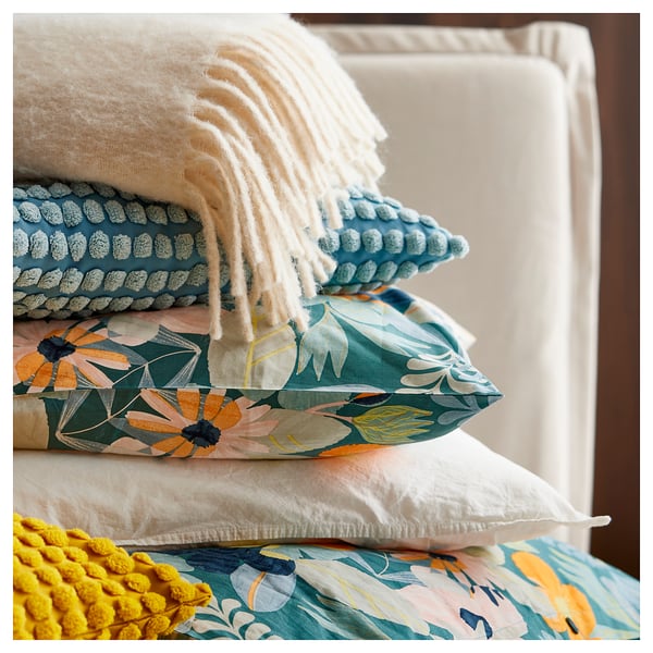 Bed met kleurrijke bloemenkussens, waaronder groenblauw, oranje en wit. Een witte gebreide plaid en een gele getextureerde deken zijn ook zichtbaar.