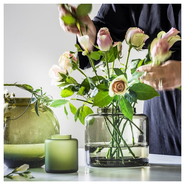 Een persoon die lichtroze rozen in een doorzichtige KONSTFULL-vaas plaatst, met twee andere vazen, een groene en een grotere, op een tafel.