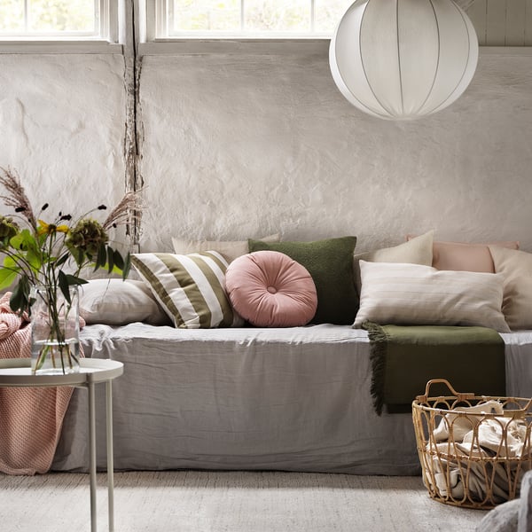 Gezellige woonkamer met grijs KRANSBORRE rond fluwelen kussen op wit linnen bank, groene plaid, en gevlochten mand op beige tapijt.