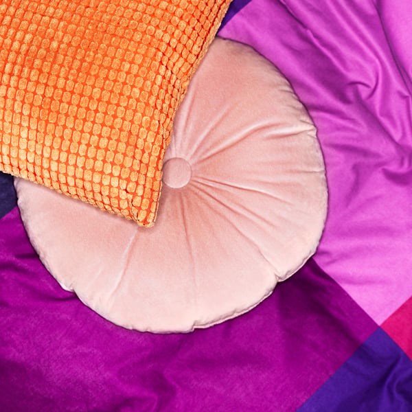 Heldere sierkussens, waaronder een rond lichtroze kussen en een kussen met oranje patroon, tegen een paarse achtergrond.
