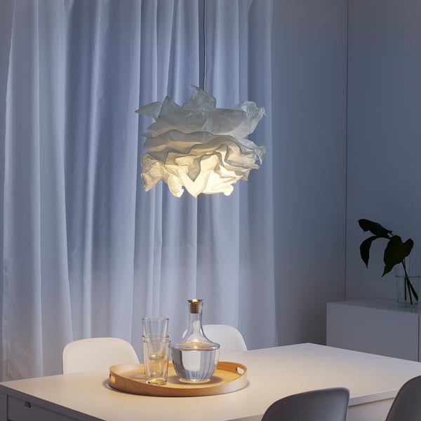 KRUSNING lamp boven eettafel met glazen en vaas, zacht wit licht, omgeven door stoelen, raam omlijst door gordijnen.