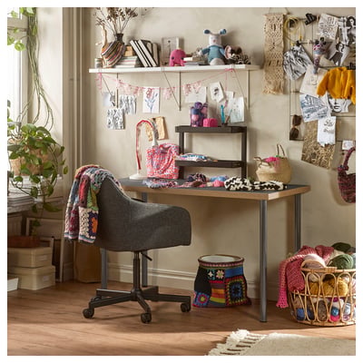 Knutselkamer met LAGKAPTEN bureau, planken met garen, gebreide artikelen en decoratieve voorwerpen. Grijze stoel-, plant- en wanddecoraties.