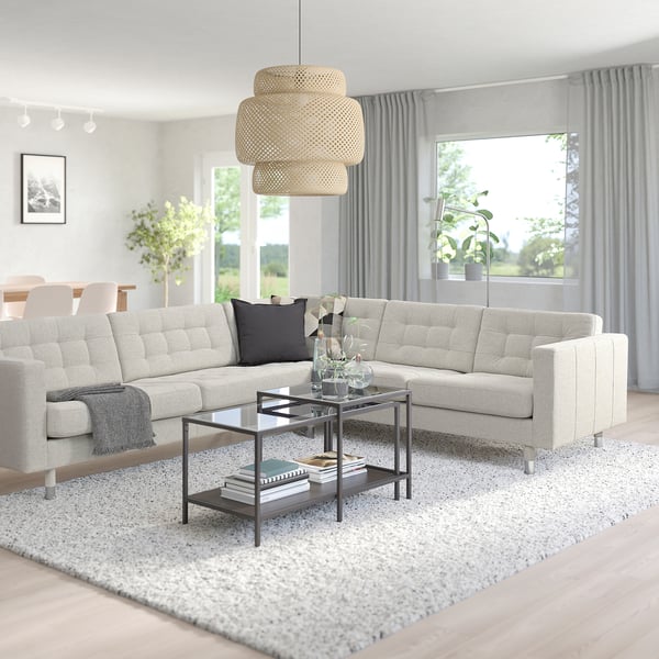 Moderne woonkamer met landnrona beige hoekbank, twee kussens, salontafel met glazen blad, groot raam en hanglamp.