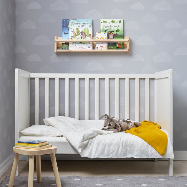 Lichte kinderkamer met wit peuterbed, knuffel, boeken en wandplank. Grijze en witte muur met wolkenpatroon. Gele deken voegt kleur toe.