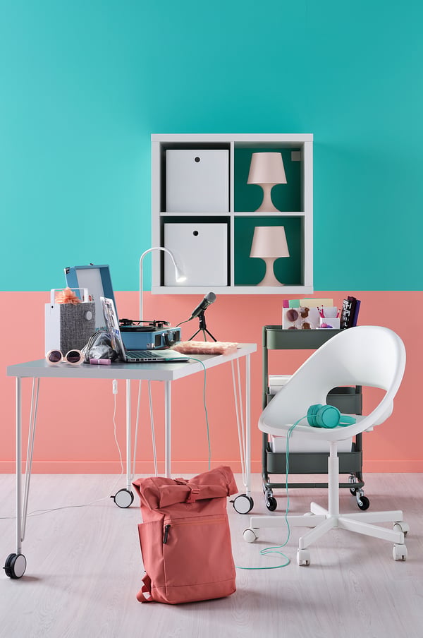 Lichte, moderne studieopstelling. Wit bureau met laptop, roze lampen en rugzak. Groene muur, groenblauwe accenten.