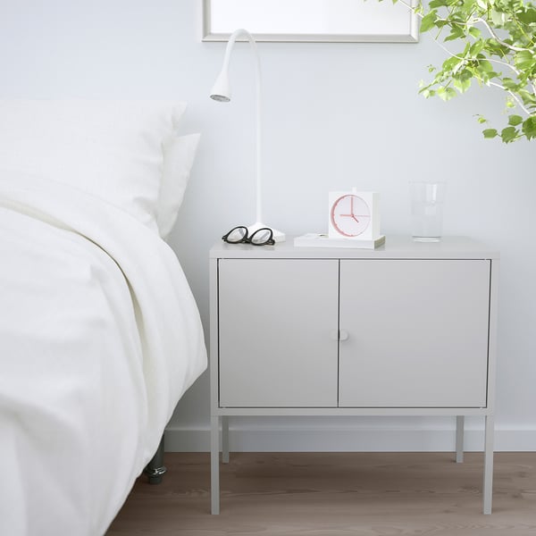 Een witte IKEA LIXHULT opbergkast naast een bed. Het is compact, met twee deuren, en ondersteunt een lamp, klok, glazen en een vaas.