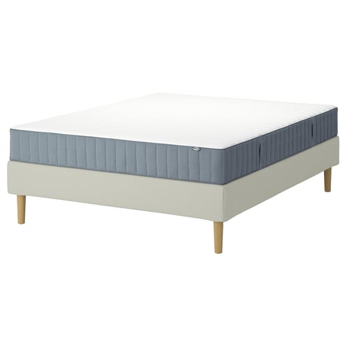Een IKEA LYNGÖR bedbasis met een wit Valevåg-matras en beige bekleding. Het bed heeft lichte houten poten en een eenvoudig, modern ontwerp.