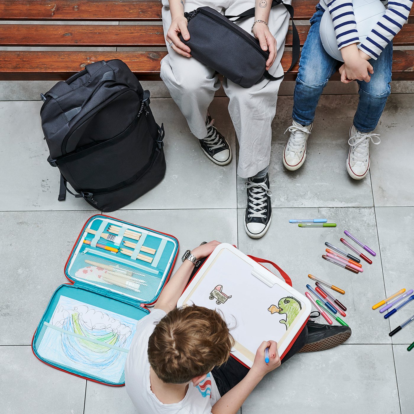 Kinderen op bankje, één tekening met open MÅLA kunstkoffer. Kleurrijke voorraden en kunstwerken op de grond, grote rugzak naast hen.
