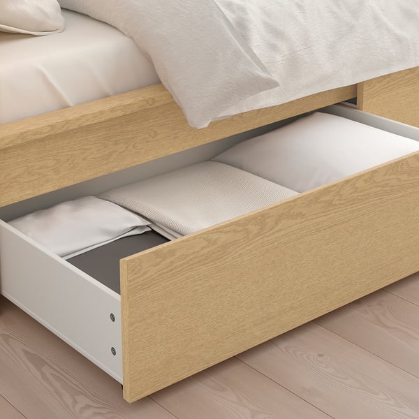 Een licht houten bed met twee grote opberglades die open kunnen en dekens binnenin onthullen. De lades zijn voorzien van zwenkwielen voor soepel rollen.