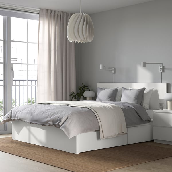 Moderne slaapkamer met wit MALM-bed, groot raam, grijze gordijnen, zachte verlichting en plant. Bed heeft lades voor opslag.