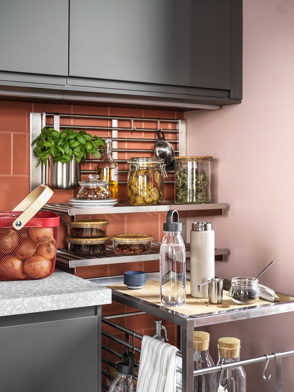 Moderne keuken met grijze kasten, roze muren en open planken met potten, kruiden, fruit en apparaten, met led-onderkastverlichting.