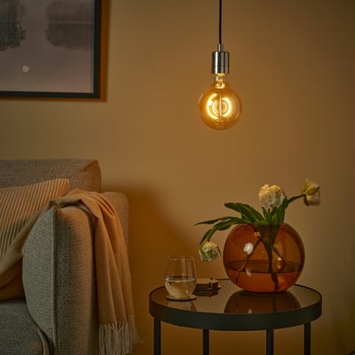 Gezellige kamer met MOLNART lamp, rode vaas & bloemen, glas, beige deken, ingelijste kunst, warm licht.