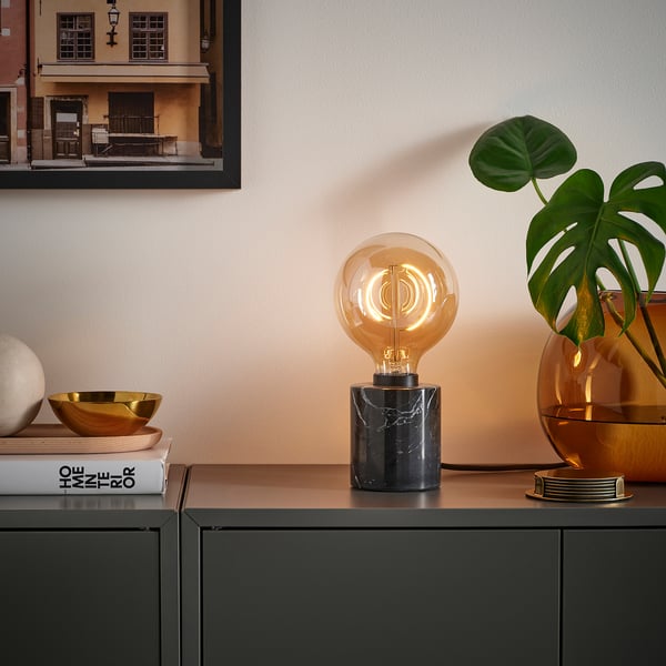 Stijlvolle lamp MOLNART met uniek filament, donkere console, boeken, gouden schaal en groene plant in warme opstelling.