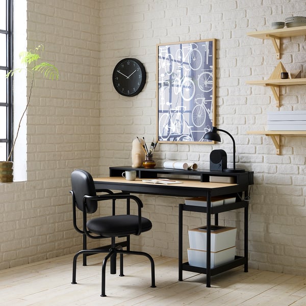 Modern zwart MULLSJÖ bureau tegen witte bakstenen muur, inclusief zwarte stoel, houten bureau met opbergruimte en unieke wandklok.