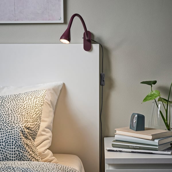 Marronrijke NÄVLINGE wandlamp bij het bed. Slank ontwerp met verstelbare arm, naast een stapel boeken en planten.