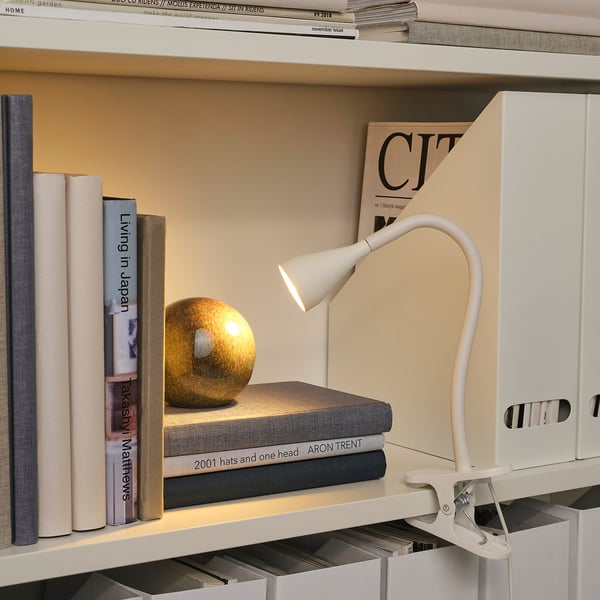 Plank met boeken & tijdschrift, verlicht door een witte, verstelbare klemlamp. Een klein boldecor rust bovenop boeken.
