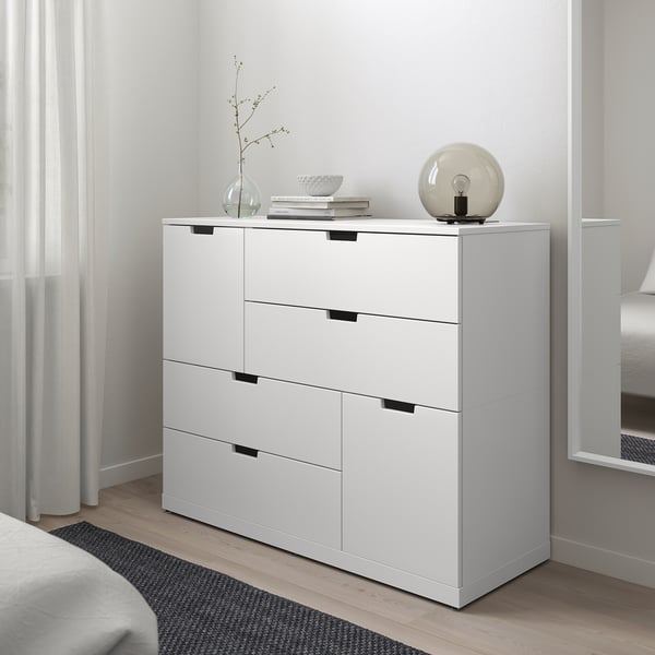 Witte NORDLI ladekast met 6 lades in de slaapkamer tussen raam en bed, moderne minimalistische stijl.