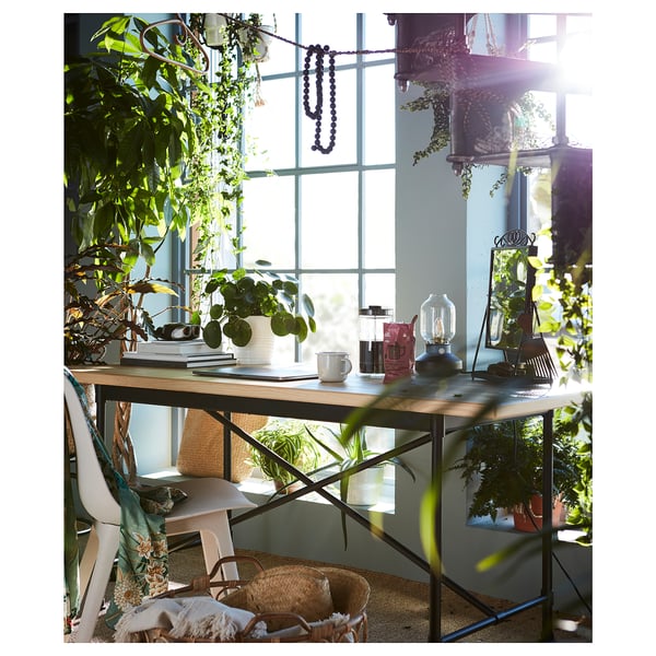Lichte, zonnige kamer met een houten bureau bij een groot raam. Bureau bevat planten, koffiezetapparaat en boeken. Gevlochten mand en witte stoel in de buurt.