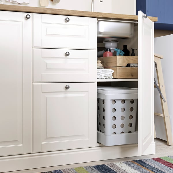 Keukenkast met geopende deur onthult opbergruimte: wasmand, opgevouwen handdoeken, zeepdispenser en houten kist.