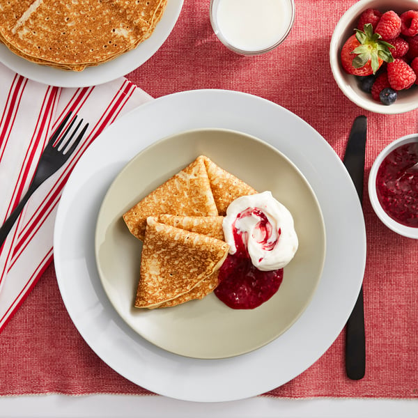 Plaat van gouden pannenkoeken gegarneerd met room en bessensaus op een rood tafelkleed.