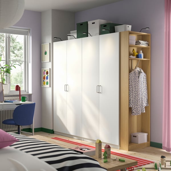 Opgeruimde slaapkamer met witte PAX-kledingkast , planken, een groene doos, hangend overhemd en een gestreepte sprei. Blauwe stoel en decor bij bureau, verlicht door een raam.