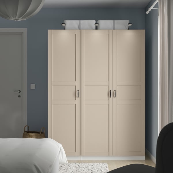 Een beige PAX kledingkast in een slaapkamer. Het is lang en breed, met vier deuren en gaasmanden erop, en staat naast een wit bed.