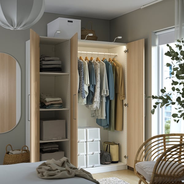 Moderne kledingkast met open deuren met kleding, opgevouwen kleding en opbergdozen. Het staat in een lichte kamer met een spiegel en een rieten stoel in de buurt.