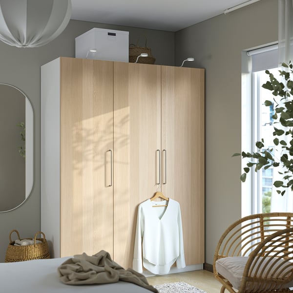 PAX kledingkast: hoge houtkleurige kast met spiegels en hangend shirt. Naast rieten stoel & plant.