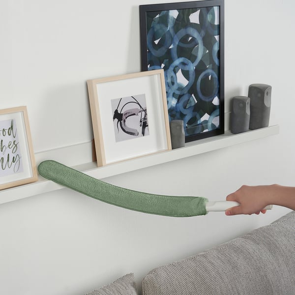Een hand die een groene PEPPRIG-microvezelstofdoekvulling vasthoudt en deze horizontaal uitrekt boven een witte plank met wanddecoratie.