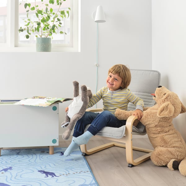 Kind zittend in een mini POÄNG fauteuil met een knuffeldier, naast een tafel met boeken in een gezellige kamer.