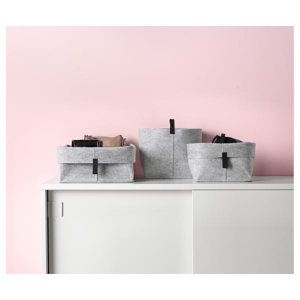 Drie lichtgrijze vilten opbergmanden op witte plank tegen roze muur, verschillende maten, gevouwen randen.
