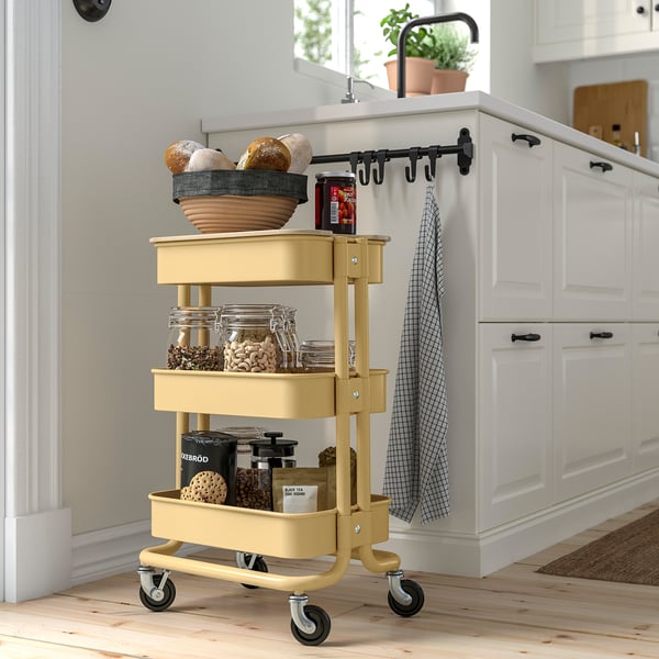 Gele RÅSKOG keukentrolley met 3 planken. Bevat brood, kruiken en potten. Compact, staal, met houten mand en wielen.