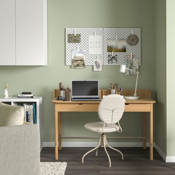 Modern thuiskantoor: licht houten bureau, witte stoel, groene muur; ophangbord, laptop, lamp.