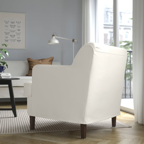 Een lichte, moderne woonkamer met een witte ROCKSJÖN-fauteuil met houten poten en een schuine rugleuning voor comfort. Grote ramen bieden uitzicht op de stad.