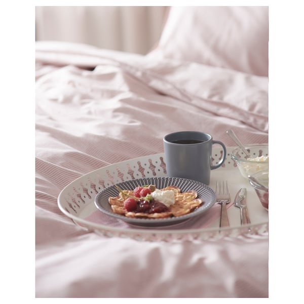 Dienblad met ontbijtartikelen, blauwe mok en bestek op roze beddengoed. Blauwe plaat met pannenkoeken, bessen en room.