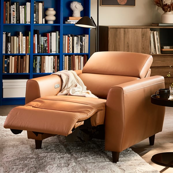 Bruin leren RULLERUM relaxstoel in een gezellige woonkamer, omringd door boekenkasten. Comfortabel en ruim, ideaal voor ontspanning.