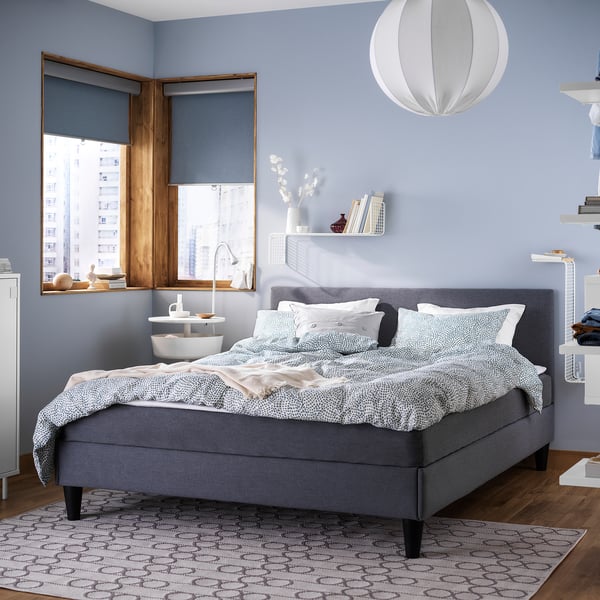 Moderne slaapkamer met grijs SÄBÖVIK-bed, blauwe muren, witte hanglamp, houten vloer en raam met uitzicht op de stad.