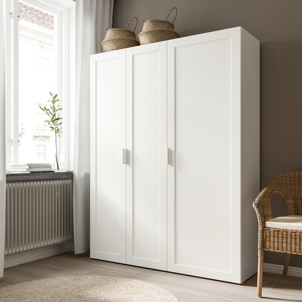 Moderne witte kledingkast met twee deuren en gevlochten manden erop, naast een raam met een radiator en plant.