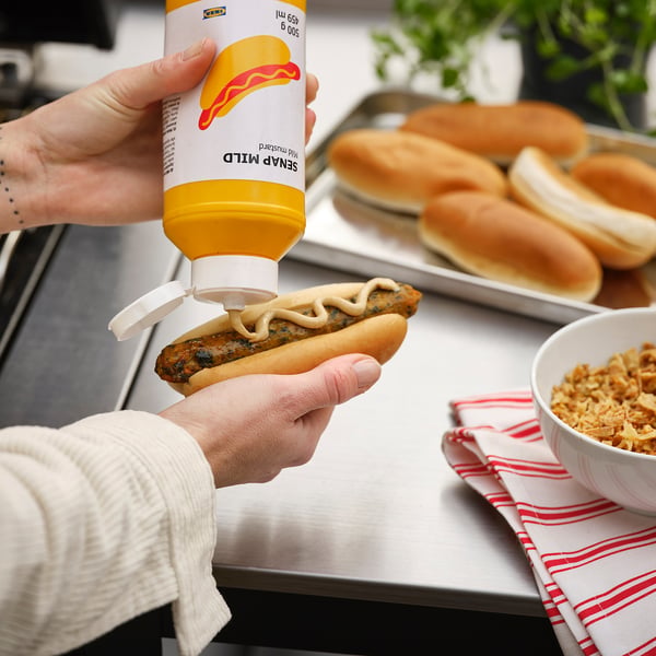 Een persoon perst gele mosterd op een hotdog. Op het etiket van de fles staat SENAP MILD.