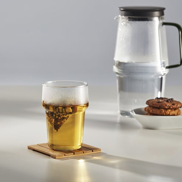 Een glazen beker op een onderzetter bevat thee met een zetgroep, naast een waterkan en koekjes op een witte tafel.