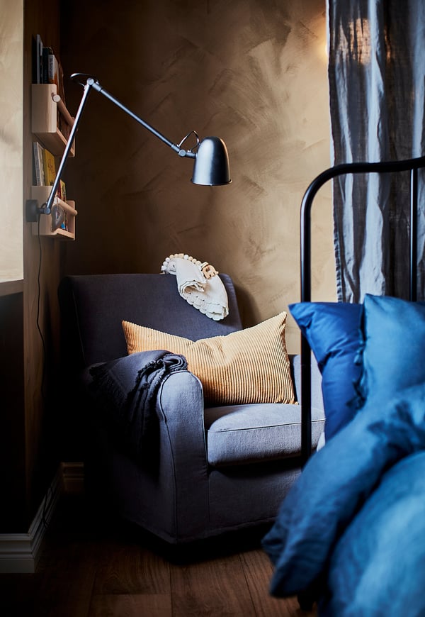 Gezellige leeshoek met grijze SKURUP-lamp, arm past zich aan, op muur naast blauwe kussens, grijze stoel, houten plank met boeken, getextureerde beige muur.