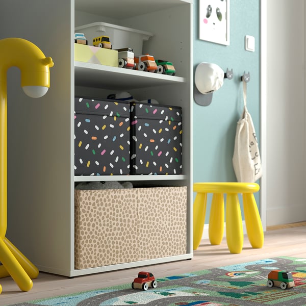 Kinderkamer met kleurrijke speelgoedopslag: SLÄKT-doos met gevlekte patroon bevat speelgoed. Felgele kruk en lamp ernaast.