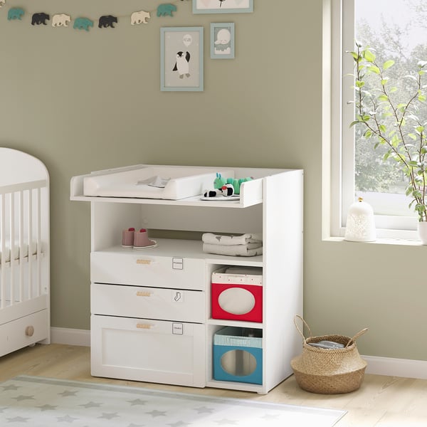 In een kinderkamer staan op een witte kinderkamertafel met planken en lades babyartikelen. Versierd met speelgoed en boeken, staat het bij een raam met planten.