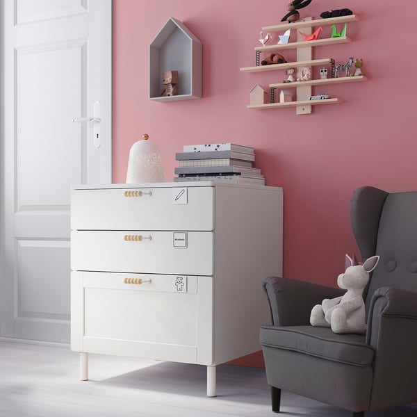 Kleine kinderkamer met witte opbergkast, grijze stoel en roze modellen op planken.