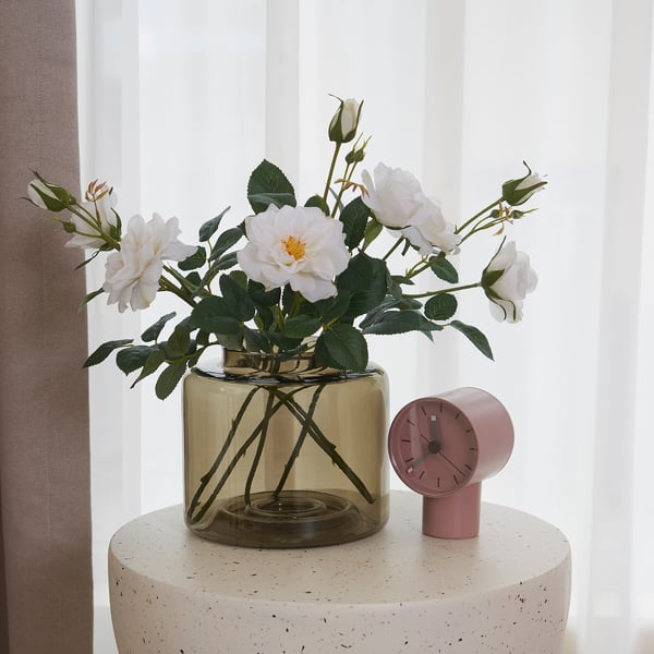 Een ronde glazen vaas met witte rozen zit op een getextureerde kruk naast een kleine roze klok.