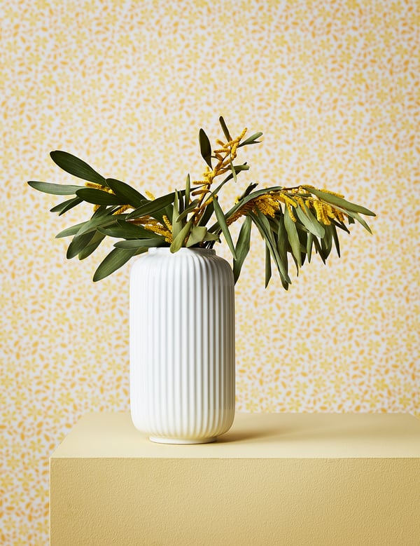 Een witte geribbelde keramische vaas, met daarin groene en gele bloemen, staat op een geel oppervlak tegen een muur met een geel patroon.