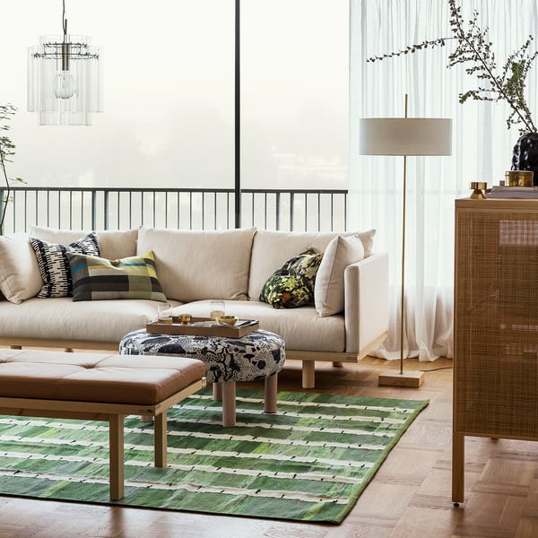 Moderne woonkamer met beige bank, vloerkleed met groen patroon, stijlvolle vloerlamp met stoffen kap en messing voet naast groot raam.