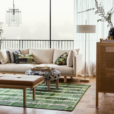 Moderne woonkamer met beige bank, vloerkleed met groen patroon, stijlvolle vloerlamp met stoffen kap en messing voet naast groot raam.