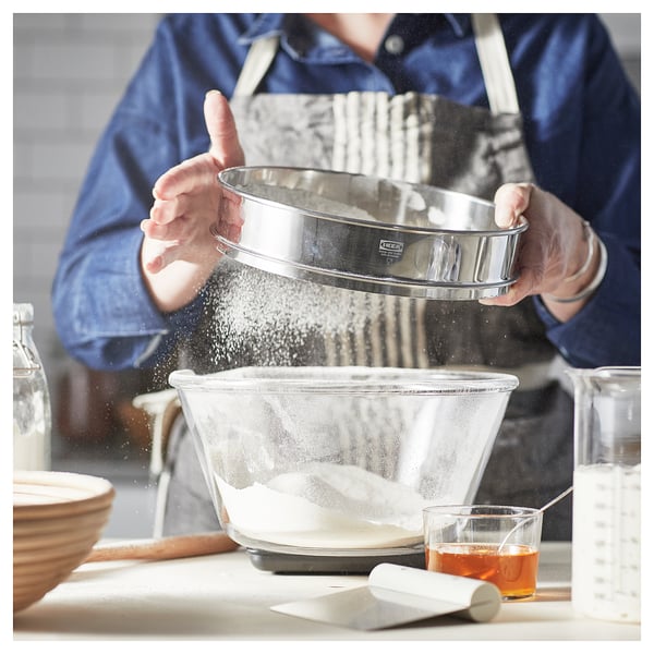 Een persoon zeeft bloem met behulp van een STRÖAD-zeef in een kom, omringd door bakgereedschap in een gezellige keuken.